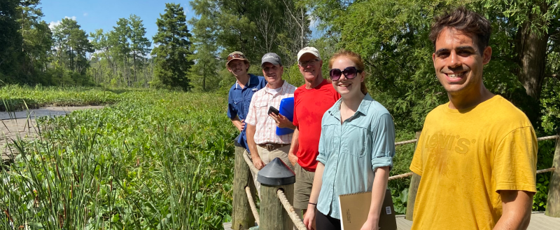Four students and a professor smiling in a row on a field trip to Roosevelt Island. A landscape of woods and marsh is visible behind them.